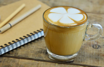 Close-up of frothy coffee by book and pen on wooden table