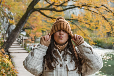 Portrait of beautiful woman pulling hat down over eyes, standing on path in part in autumn