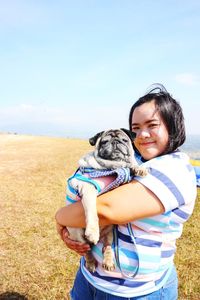 Portrait of woman holding dog while standing on field against blue sky