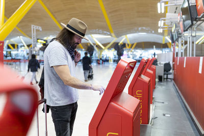 Young man with beard and long hair and hat buying airline tickets at the airline's automatic terminal