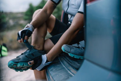 Cropped unrecognizable female bicyclist in helmet and sunglasses sitting in trunk of car and preparing for ride while putting on boots and looking at camera