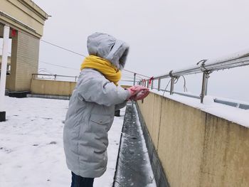 Girl in warm clothing playing with snow on terrace against sky
