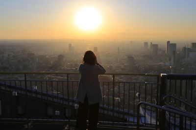 Rear view of woman standing by railing against sky during sunset