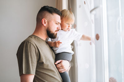 Multinational family, young man father with baby girl on window sill looking at window at home