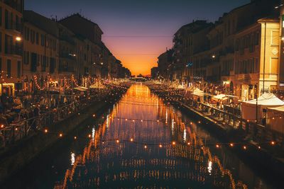 Illuminated street by buildings against sky at night