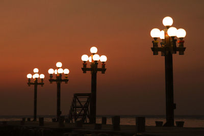Illuminated street light by sea against sky at sunset