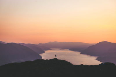 Mid distance of man standing on cliff by lake against sky during sunset