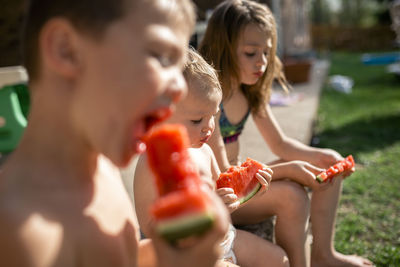 Siblings eating watermelons while sitting at yard