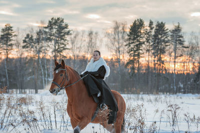 A girl in a white cloak rides a brown horse in winter.