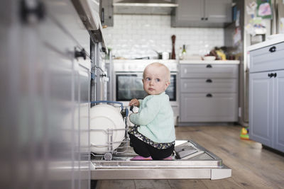 Cute girl looking away while sitting in dishwasher at kitchen