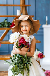 Portrait of girl wearing hat while standing with flowers against wooden steps at home
