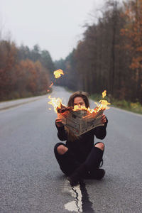 Young woman sitting on the empty road, holding burning paper in her hands