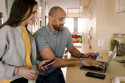 Smiling girl looking at father doing online shopping while sitting at kitchen island