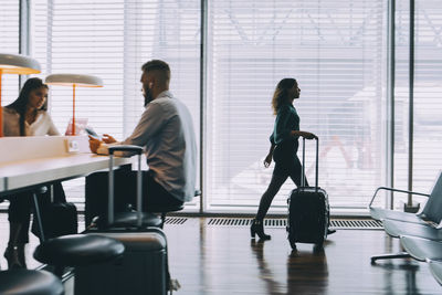 Full length of businesswoman walking with luggage by window at airport departure area