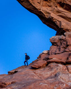 Low angle view of man standing on rock against clear blue sky