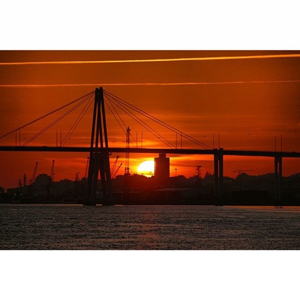 sunset, built structure, orange color, architecture, silhouette, connection, sky, sun, building exterior, bridge - man made structure, suspension bridge, water, engineering, clear sky, bridge, sea, river, city, outdoors, no people