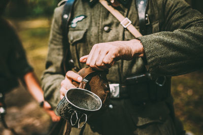 Midsection of army soldier holding bottle and mug while standing in forest