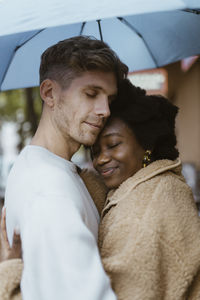 Side view of multiracial romantic couple embracing with eyes closed under umbrella