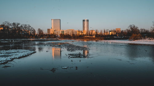 Reflection of buildings in lake against sky in city during winter