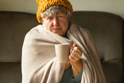 Elderly woman in ginger cap wrapped in blanket and drinking cup of tea. grandma feeling cold at home