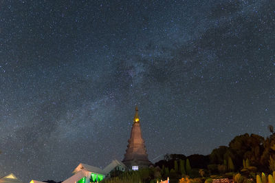 Low angle view of cross amidst buildings against sky at night milky way galaxy