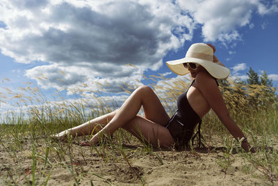 A woman in a swimsuit, hat and sunglasses sunbathes in summer on the riverbank among the grass