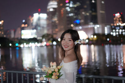 Portrait of young woman standing against illuminated city at night