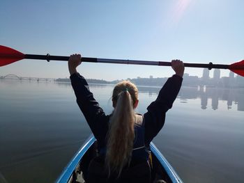 Rear view of mid adult woman holding oar with arms raised while sitting in canoe on lake