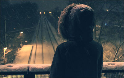 Rear view of person standing on footbridge over snow covered railroad tracks at night