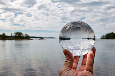 Cropped image of hand holding crystal ball against lake and cloudy sky