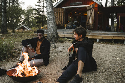 Male friends with drinking glass talking while sitting by fire pit during weekend
