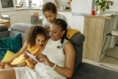 Happy woman sharing smart phone with daughter and son on sofa at home