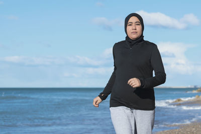 Portrait of young woman standing at beach against sky