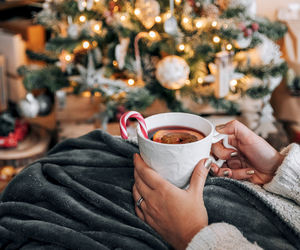 Close-up of woman in warm sweater holding cup of hot beverage in front of christmas tree.