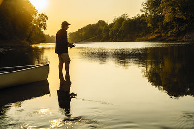 Side view of mid adult man fishing while standing in lake during sunset