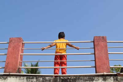 Rear view of boy on railing against clear blue sky