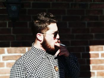 Young man looking away while smoking cigarette against brick wall