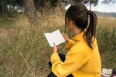 Side view of unrecognizable female sitting in filed in autumn and enjoying interesting story in book