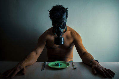 Portrait of young man wearing mask sitting on table against wall