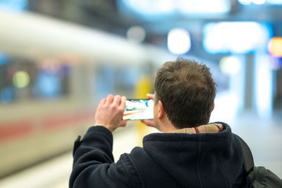 Rear view of man photographing with phone at railroad station platform