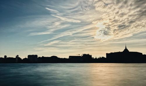 Silhouette buildings by river against sky during sunset