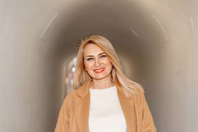 A cheerful blonde woman in a warm camel coat stands in an urban tunnel, radiating joy