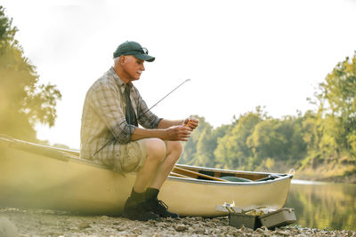 Senior man adjusting fishing tackle while sitting on boat at lakeshore against clear sky