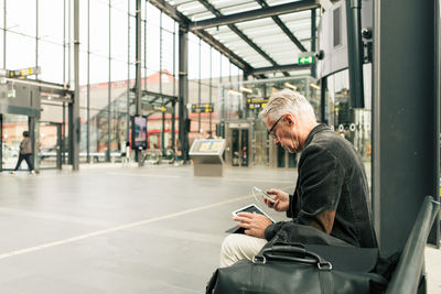 Senior male commuter sitting with technologies and bags at railroad station