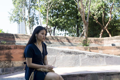 Young woman looking away while sitting on retaining wall against trees