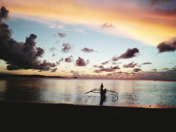 Silhouette man standing on boat with outrigger in sea against sky during sunset