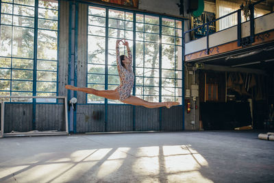 Side view of young slim ballerina in gymnastic suit jumping while practicing dance movements in light spacious studio with large window