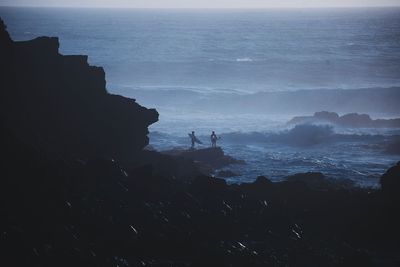 Silhouette people on rocks at beach against sky