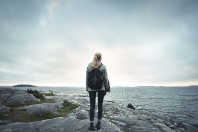 Rear view of woman standing on rock by sea against cloudy sky