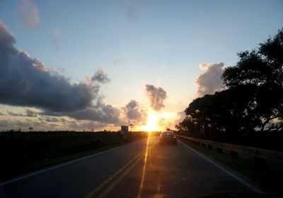 Road by trees against sky during sunset